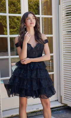 A woman stands before glass doors in the Nox Anabel R877—a chic black short ruffled lace off-the-shoulder homecoming dress—her hand gracefully touching her hair.