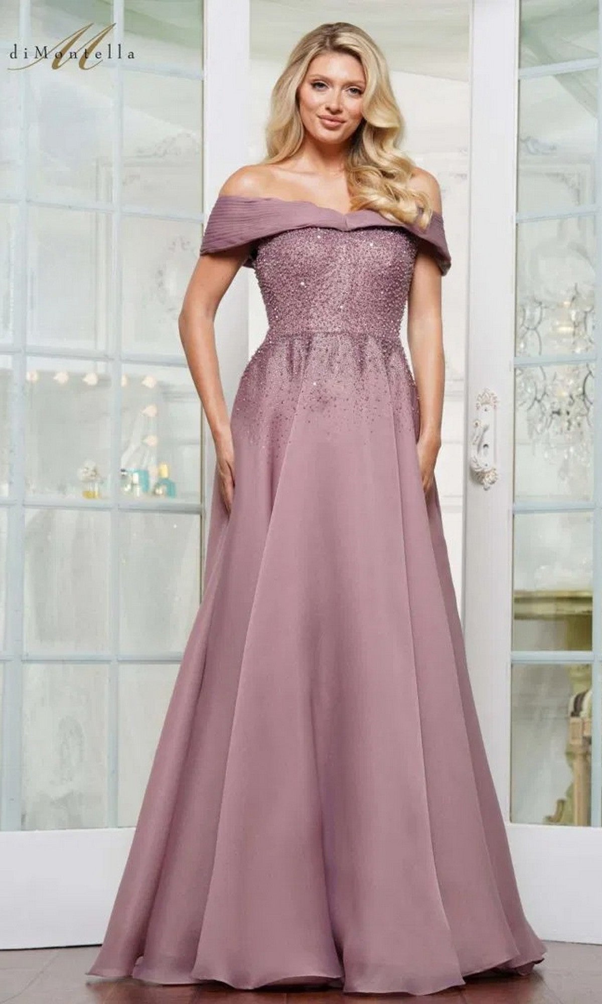 A woman stands indoors wearing the Rina Di Montella RD3102 long formal dress in Victorian Lilac, featuring an off-the-shoulder beaded bodice and a flowing, floor-length A-line skirt.