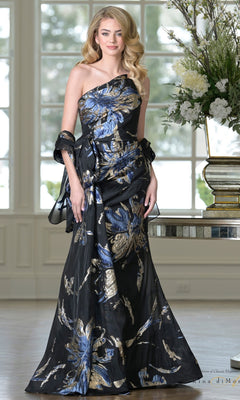 A woman stands indoors in the Rina Di Montella RD3233, a one-shoulder formal gown—floor-length, black with blue and gold floral patterns—paired with a matching wrap.