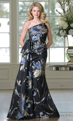 A woman smiles indoors, hands in the pockets of the Rina Di Montella RD3233 one-shoulder formal gown, featuring a black base with bold blue and gold metallic floral print.