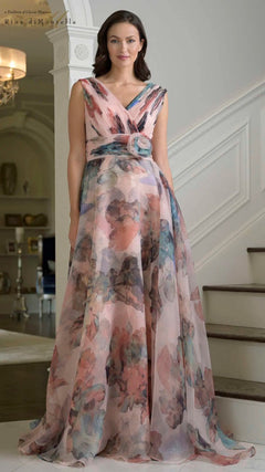 A woman stands indoors on a staircase wearing the Rina Di Montella RD3426 long formal dress in Mauve Multi, featuring a sleeveless organza design with a floral-patterned draped bodice and flower detail at the waist.