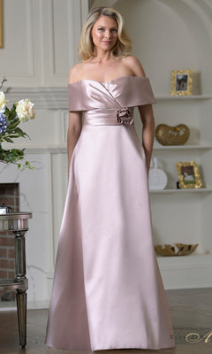 A woman stands in a decorated room wearing the Rina Di Montella RD3429 long formal dress, featuring an off-the-shoulder design, light pink color, and a floral detail at the waist.
