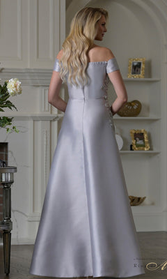 A woman with long blonde hair stands facing away in an elegant room, wearing the Rina Di Montella RD3501—an off-the-shoulder, floor-length silver Mikado formal dress with an a-line skirt.