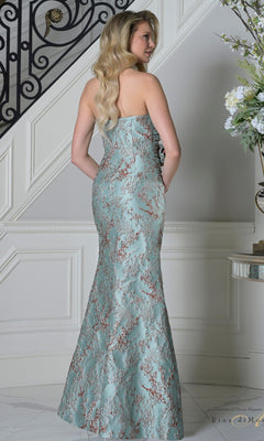 A woman wearing the Rina Di Montella RD3502 long formal dress, featuring a strapless jacquard design with a gold floral pattern and floor-length skirt, stands on an indoor staircase facing away from the camera.