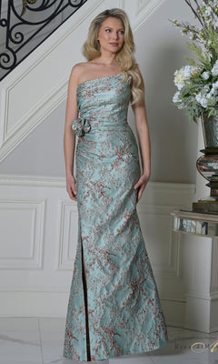 A woman wearing the Long Formal Dress: Rina Di Montella RD3502 in Sage Crimson stands indoors near a staircase and floral arrangement.