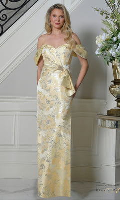 A woman stands indoors wearing the Rina Di Montella RD3503 long formal dress in canary—a floor-length gown with a sweetheart neckline, gold and silver floral texture, off-the-shoulder design, and an elegant side bow detail.