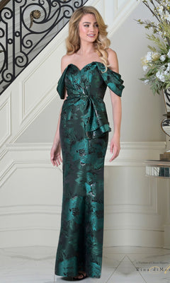 A woman stands indoors, smiling in the jade Long Formal Dress: Rina Di Montella RD3503—an off-the-shoulder evening gown with a sweetheart neckline, floral jacquard fabric, and a bow at the waist—near a staircase and floral arrangement.