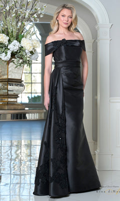 A woman stands indoors beside a white pillar and large flower arrangement, wearing the black Rina Di Montella RD3504 Long Formal Dress with floral lace details, off-the-shoulder neckline, and ruched waistband.