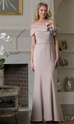 A woman wearing the Rina Di Montella RD3507 long formal dress in Champagne Gd stands in a tastefully decorated room with a fireplace and shelves in the background.