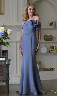 A woman in a Rina Di Montella RD3507 deep blue, off-the-shoulder, floor-length formal dress with floral detailing stands in a living room with decorative shelves and a fireplace.