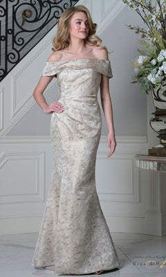 A woman in a champagne Long Formal Dress: Rina Di Montella RD3511 with an off-the-shoulder neckline and floor-length metallic brocade stands indoors beside a staircase and floral arrangement.