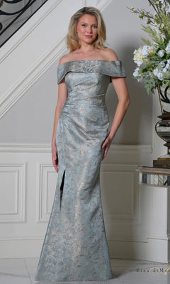 A woman in a deep sage Rina Di Montella RD3511 long formal dress with an off-the-shoulder neckline and front slit stands indoors near a staircase and floral arrangement.
