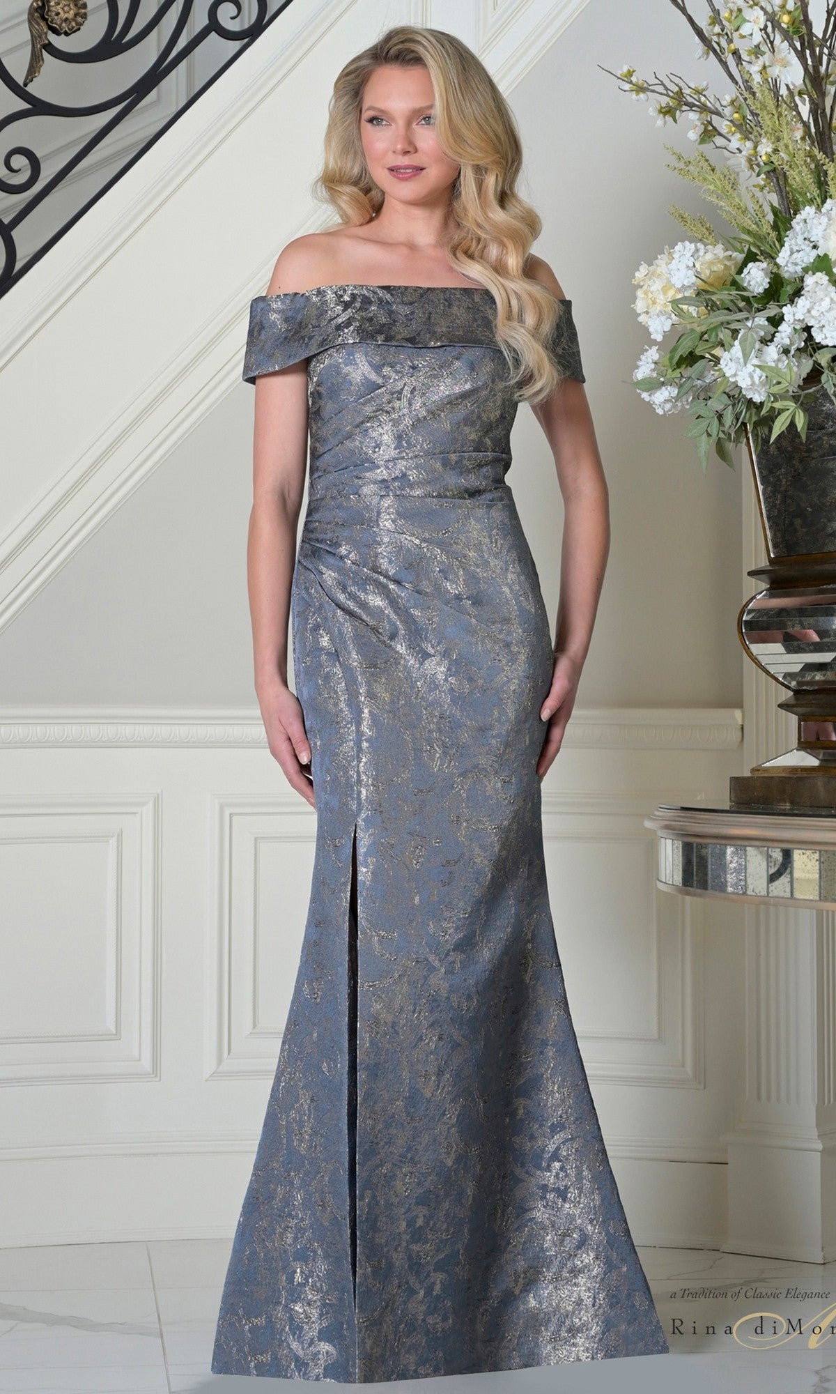 A woman stands indoors near a staircase and floral arrangement, wearing the Rina Di Montella RD3511 long formal dress in Wedgewood—a floor-length gown with an off-the-shoulder neckline and subtle metallic brocade pattern.