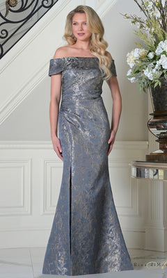 A woman stands indoors near a staircase and floral arrangement, wearing the Rina Di Montella RD3511 long formal dress in Wedgewood—a floor-length gown with an off-the-shoulder neckline and subtle metallic brocade pattern.