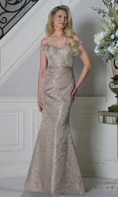 A woman in the Rina Di Montella RD3513 long formal dress in dark taupe, featuring a fitted bodice, jacquard fabric with subtle metallic pattern, off-the-shoulder neckline, and floor-length silhouette, stands by a staircase and elegant floral display—ideal for mother-of-the-bride.