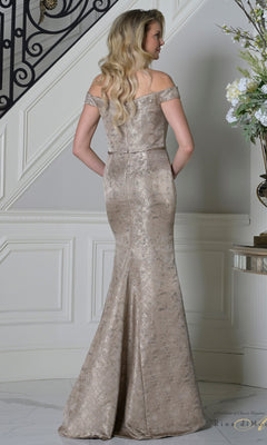 A woman with long blonde hair wears the Rina Di Montella RD3513 long formal dress in champagne, featuring an off-the-shoulder jacquard design with a subtle metallic pattern, as she stands indoors near a staircase and flowers.
