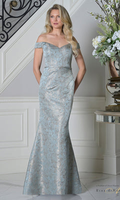 A woman wears the Rina Di Montella RD3513 long formal dress in deep sage with a metallic floral jacquard pattern, standing indoors near a staircase and flowers—an elegant option for a mother-of-the-bride.