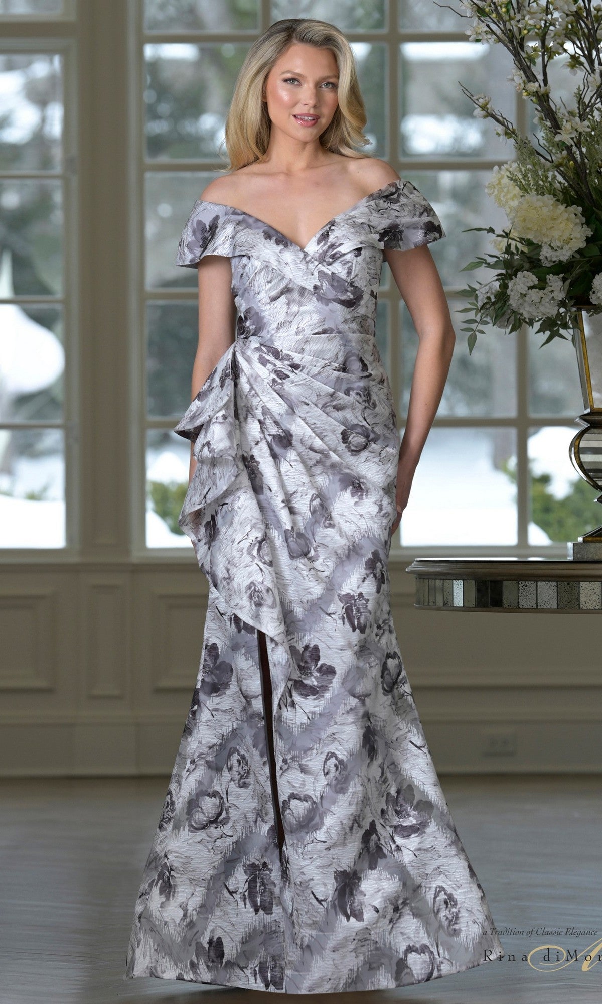 A woman stands indoors by a flower-adorned table, wearing the Rina Di Montella RD3518 long formal dress in Gray Multi with an off-shoulder neckline and mermaid skirt, as snowy scenery is visible through large windows.