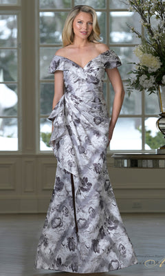 A woman stands indoors by a flower-adorned table, wearing the Rina Di Montella RD3518 long formal dress in Gray Multi with an off-shoulder neckline and mermaid skirt, as snowy scenery is visible through large windows.