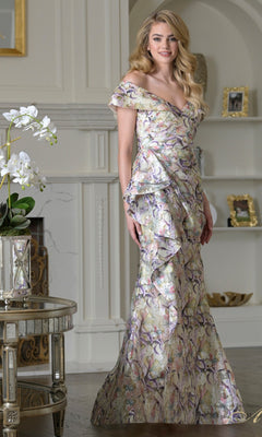 A woman stands indoors wearing the Rina Di Montella RD3518 long formal dress in Yellow Multi, an off-the-shoulder jacquard organza gown with a multicolored floral pattern and ruffled details, surrounded by flowers and framed pictures.