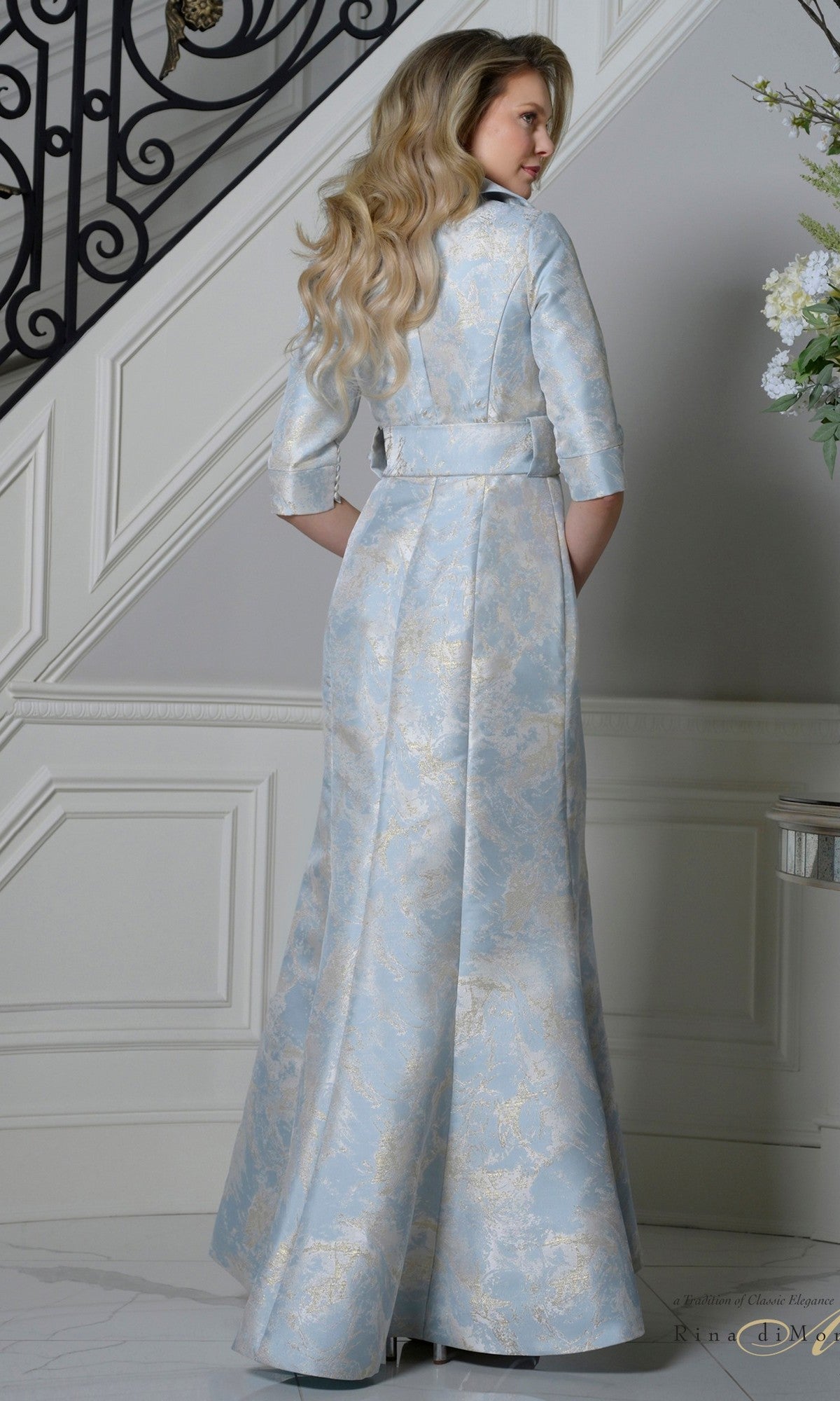 A woman with long wavy hair stands on a staircase landing, wearing the Rina Di Montella RD3521 long formal dress in light blue and silver jacquard with three-quarter sleeves, a belt, and elegant detailing.