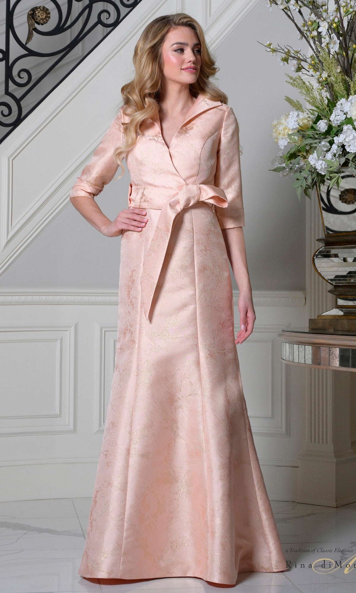 A woman stands indoors by a staircase and floral arrangement, wearing the Rina Di Montella RD3521 long formal dress in blush, featuring jacquard fabric, elbow-length sleeves, and a wide collar.
