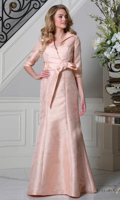 A woman stands indoors by a staircase and floral arrangement, wearing the Rina Di Montella RD3521 long formal dress in blush, featuring jacquard fabric, elbow-length sleeves, and a wide collar.