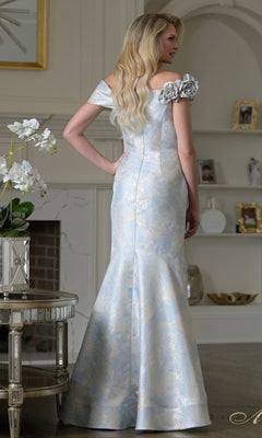 A woman with long blonde hair wears the Rina Di Montella RD3522, a light blue and silver jacquard off-the-shoulder gown with floral accents, standing in a softly lit, elegant living room.