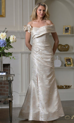 A woman stands indoors wearing the Rina Di Montella RD3522 long formal dress in champagne satin, featuring floral embellishment on one shoulder and elegant off-shoulder sleeves—perfect for any formal occasion.