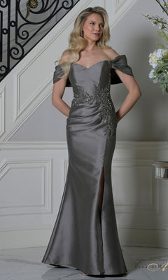 A woman indoors wears the Rina Di Montella RD3527 long formal dress in gunmetal, featuring an off-the-shoulder mermaid silhouette with floral embellishments and a front slit.