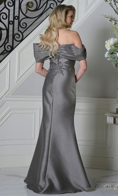 A woman with long blonde hair stands near a staircase, her back to the camera, wearing the elegant silver Rina Di Montella RD3527 long formal off-the-shoulder mermaid dress.