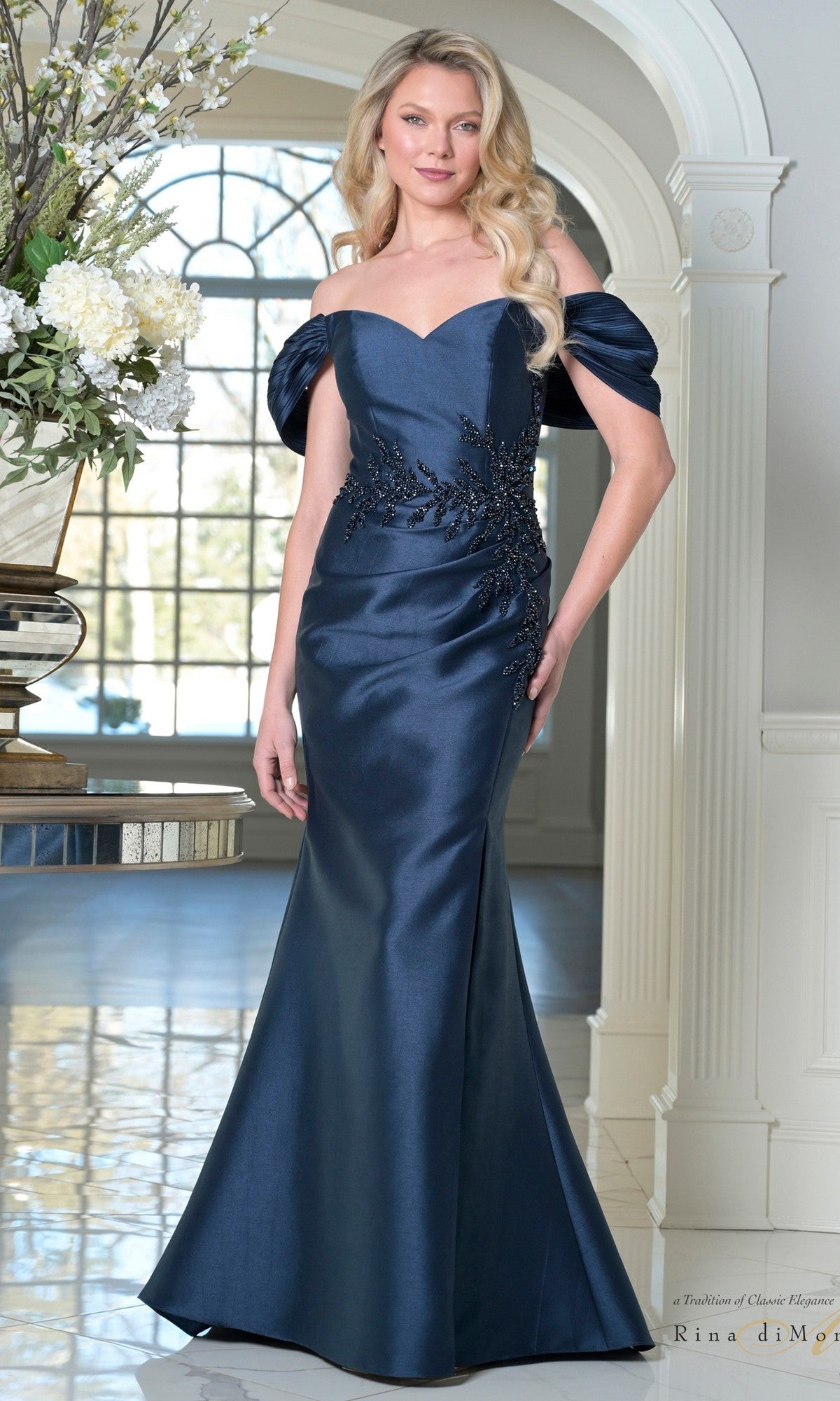 A woman in a navy Long Formal Dress: Rina Di Montella RD3527 stands indoors by a window and floral arrangement—an elegant, off-the-shoulder choice with floral appliqué for a mother of the bride.