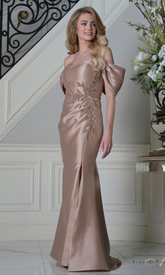 A woman stands indoors by a staircase, wearing the Rina Di Montella RD3527 long formal dress in rose gold, featuring off-the-shoulder pleated sleeves and elegant embroidery.