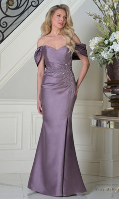 A woman stands indoors wearing the Rina Di Montella RD3527 long formal dress in Victorian, an off-the-shoulder, floor-length mermaid style with floral embroidery and draped sleeves; a staircase and flowers appear in the background.