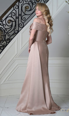A woman with long blonde hair wears the Long Formal Dress: Rina Di Montella RD3528—an off-shoulder, floor-length beige satin gown with 3D floral appliques—standing on a white marble floor before an ornate staircase.