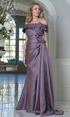 A woman stands indoors in the Rina Di Montella RD3528 long formal dress in Victorian Lilac, featuring an off-shoulder neckline and 3D floral appliques on the bodice. Marble flooring and decorative flowers are seen in the background.