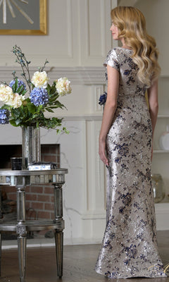 A woman in the Rina Di Montella RD3529 long formal dress stands indoors by a vase of white and blue flowers, facing away from the camera.