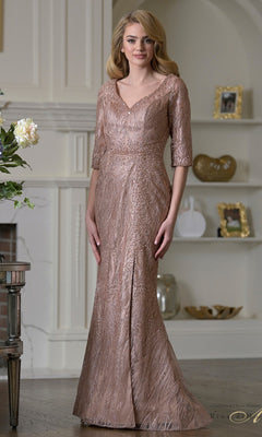 A woman wears the Long Formal Dress: Rina Di Montella RD3530 in rose gold, featuring embroidered mesh half sleeves, standing in a stylish room adorned with shelves and framed photos.