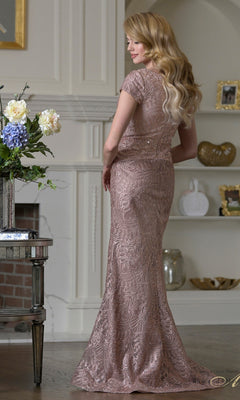 A woman with long blonde hair stands indoors, facing backward, wearing the fitted beaded Rina Di Montella RD3532 long formal dress with a mermaid silhouette. Shelves, a fireplace, and a table with flowers are in the background.