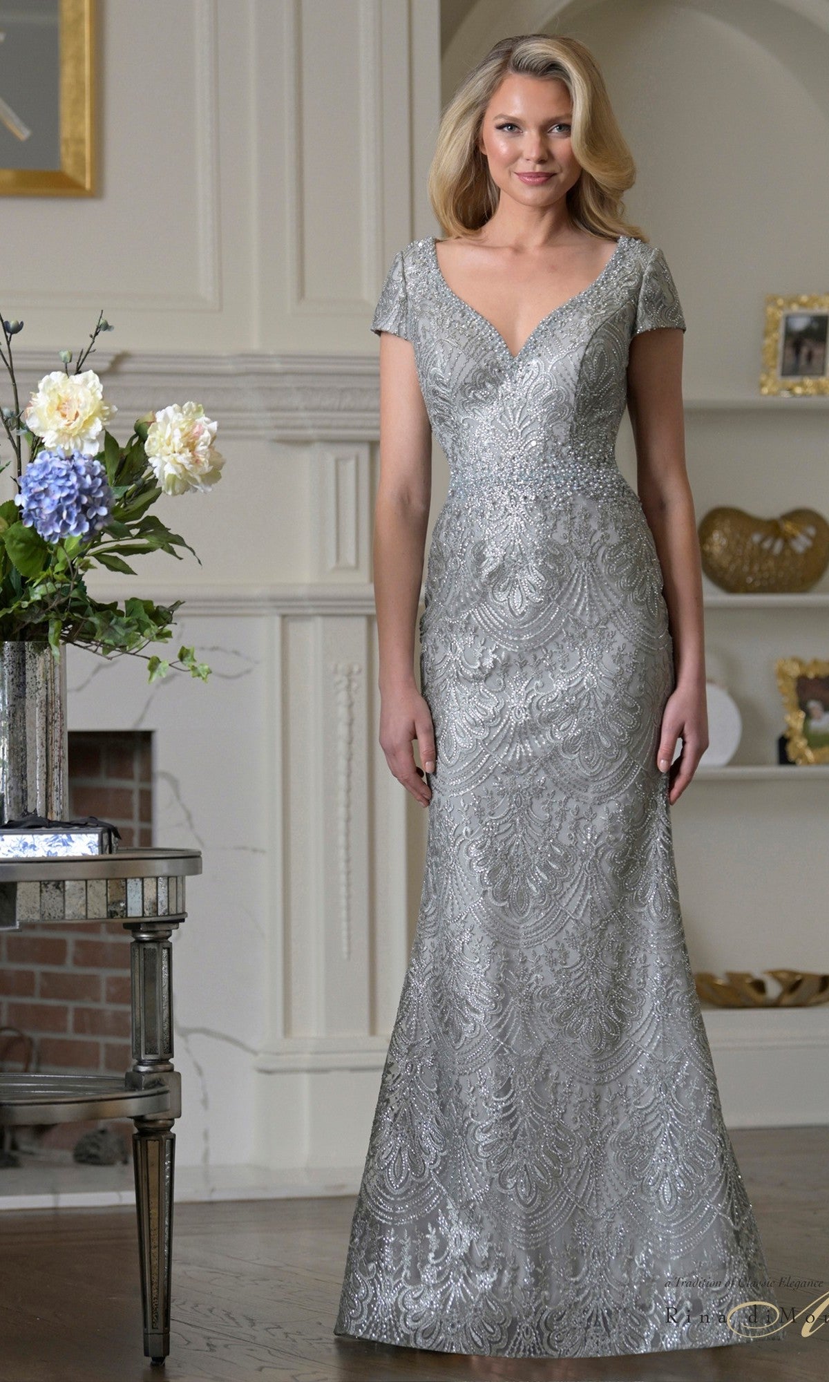 A woman poses indoors in the Rina Di Montella RD3532, a seaglass long formal dress with a fitted bodice and intricate beaded embroidery, evoking an elegant mother-of-the-bride look by the mantelpiece.