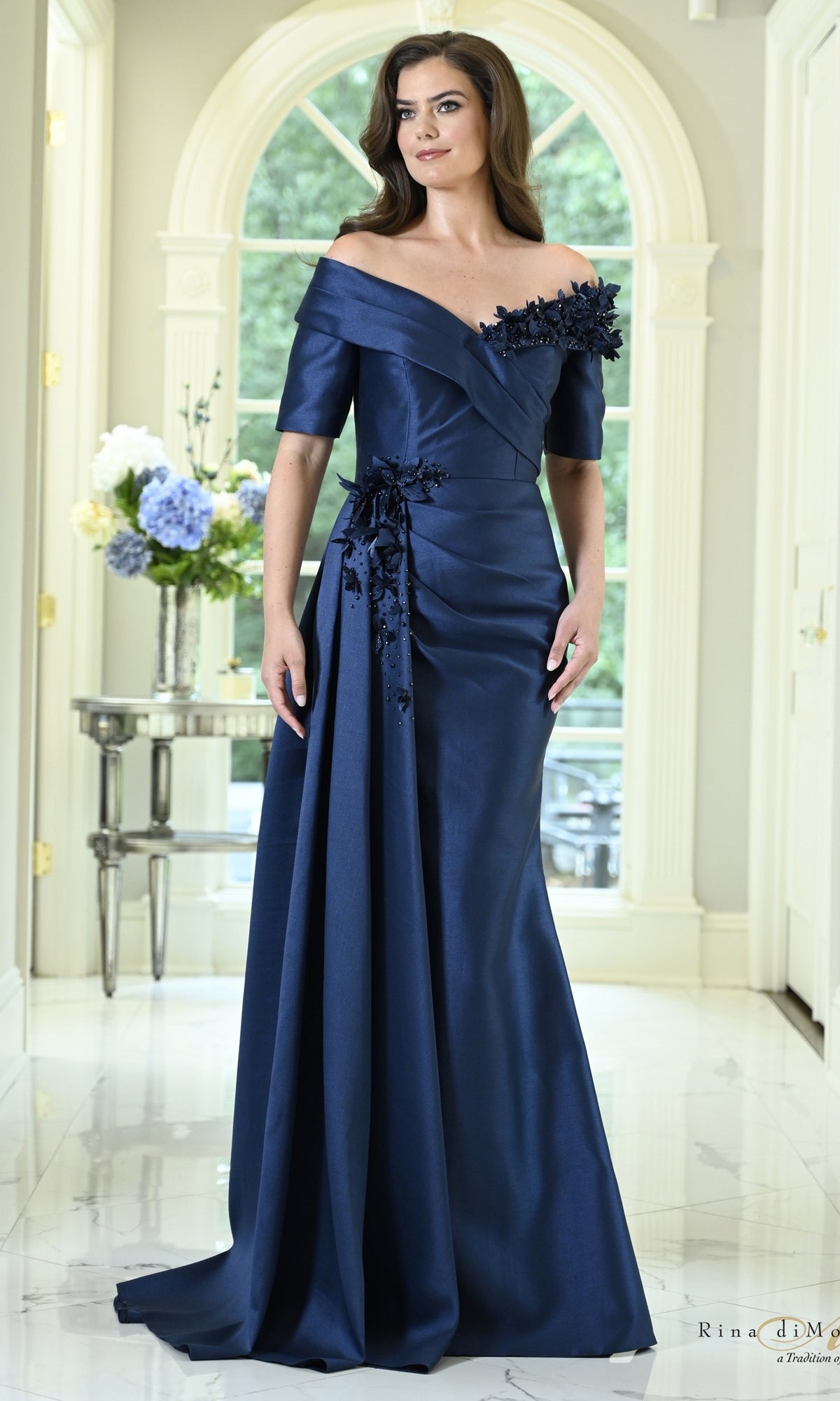 A woman wears a navy Off-the-Shoulder Floral Mermaid Formal Dress: Rina Di Montella RD3611, featuring an off-the-shoulder floral mermaid silhouette with delicate appliqué on the shoulder and hip.