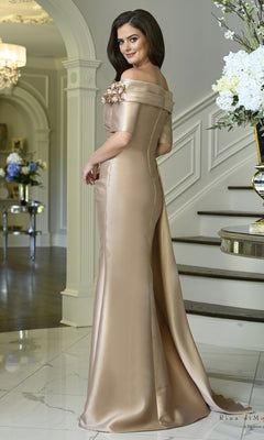 A woman stands in a foyer wearing an Off-the-Shoulder Floral Mermaid Formal Dress: Rina Di Montella RD3611, an off-the-shoulder champagne mermaid gown featuring floral detail on the bodice.