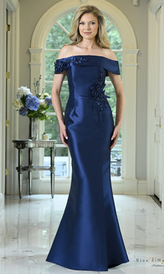 A woman wears a floor-length, off-the-shoulder navy mermaid Folded Off-the-Shoulder Mermaid Formal Dress: Rina Di Montella RD3612 with beaded floral embellishments on the neckline and hip.