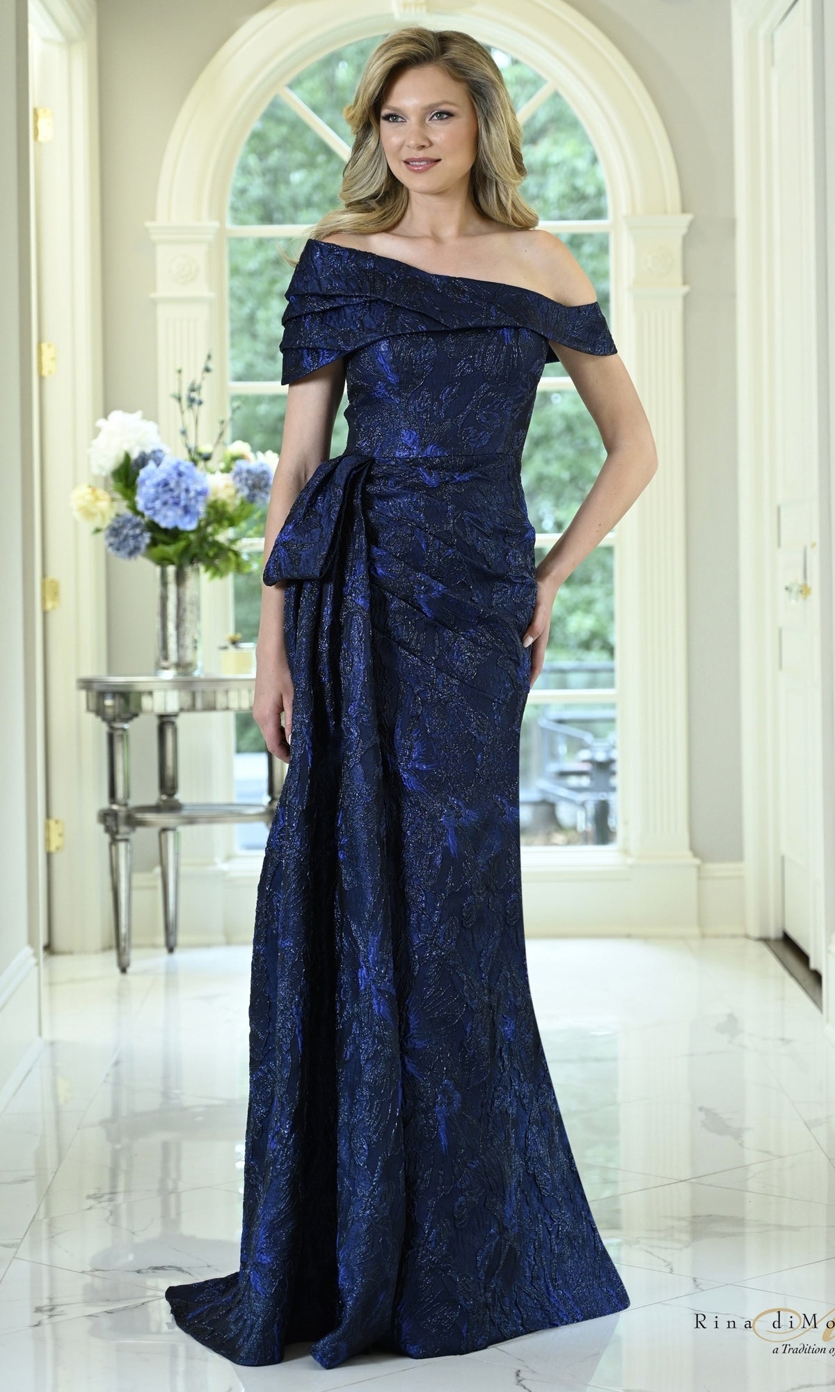 A woman wearing a navy blue Metallic Side Bow Long Formal Dress: Rina Di Montella RD3621, a floor-length, off-the-shoulder evening gown with a side bow detail at the waist.