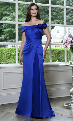 A woman wears a royal Off-the-Shoulder Rosette Satin Formal Dress: Rina Di Montella RD3639, featuring an off-the-shoulder neckline, three shoulder rosettes, and a pleated front panel on the satin gown.