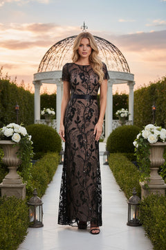 A woman wearing the R&M Richards 3253 Short Sleeve Black Formal Dress stands on a garden pathway lined with lanterns and white flowers, with a domed gazebo and sunset in the background.