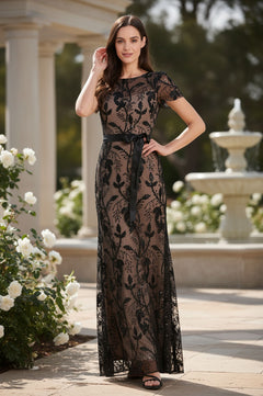 A woman stands outdoors in the R&M Richards 3253P petite long black formal dress, featuring floor-length sheer lace and a black ribbon belt, surrounded by white flowers with a stone fountain behind her.