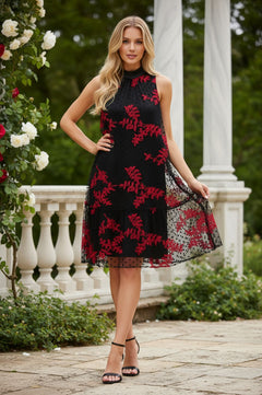 A woman poses outdoors in front of a white balustrade, surrounded by greenery and flowers, wearing the R&M Richards 4328 black/red short party dress with red floral patterns and black heels.