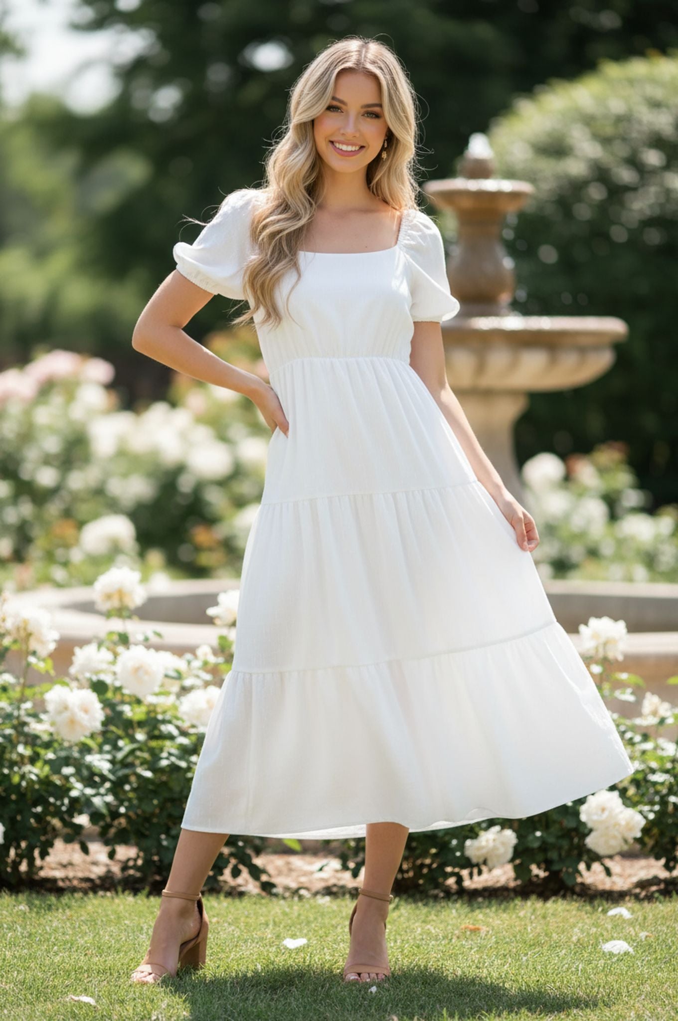 A woman stands in a garden, wearing the R&M Richards 6669 tea-length white party dress and sandals, surrounded by flowers and a fountain.