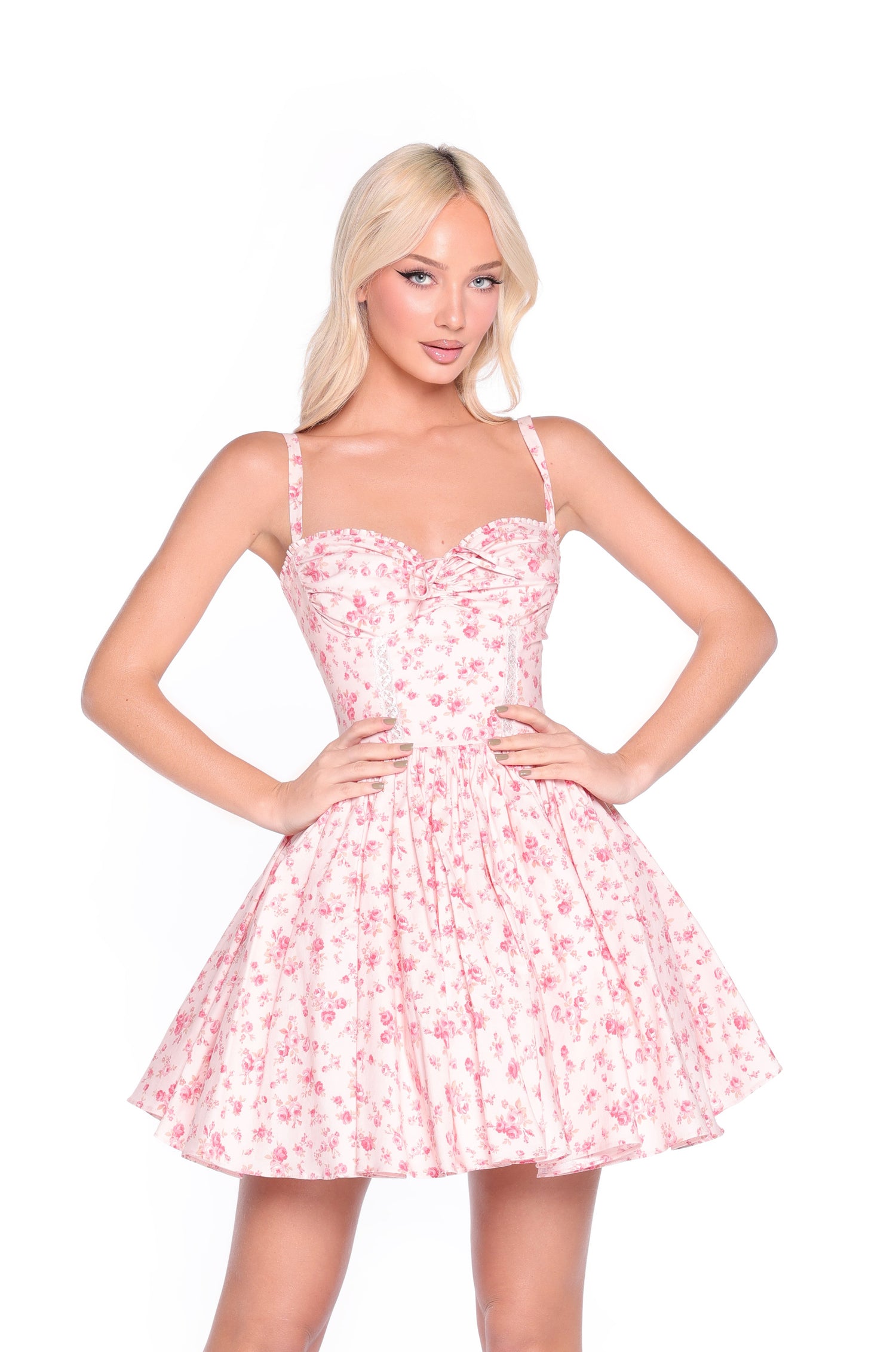 Woman wears the Blush Rose Print Floral Casual Mini Dress: Rosalind, a sleeveless fitted pink mini dress with rose print and an a-line skirt, standing hands on hips against a white background.